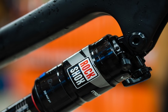 A close-up view of a black and silver RockShox mountain bike shock absorber with droplets of water on its surface. The background is a blurred orange hue, emphasizing the focus on the mechanical component.