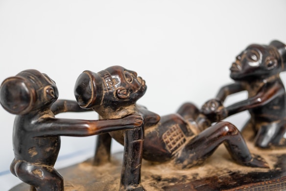 A detailed wooden sculpture depicting three stylized figures interacting in a dynamic pose. The figures have exaggerated facial features and limbs, and they are intricately carved with patterns and textures.
