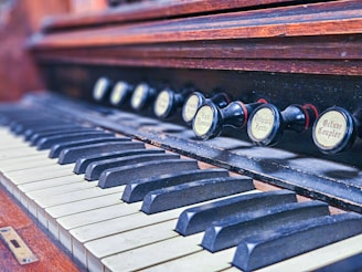 An old wooden organ keyboard with visible black and white keys. Above the keys, there are several labeled pull stops, used for controlling the sound settings of the organ. The wood appears polished, yet aged, giving it a vintage look.