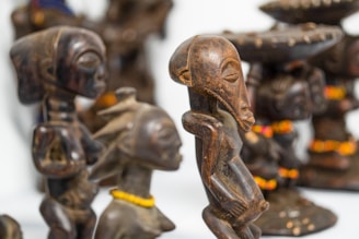 Several intricately carved wooden sculptures are displayed, featuring elongated faces and stylized bodies. The sculptures have a dark, polished finish, some adorned with colorful beaded necklaces. The craftsmanship reflects traditional art styles.