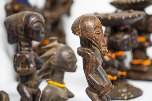 Several intricately carved wooden sculptures are displayed, featuring elongated faces and stylized bodies. The sculptures have a dark, polished finish, some adorned with colorful beaded necklaces. The craftsmanship reflects traditional art styles.