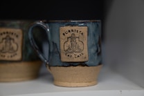 A ceramic mug with a blue glaze and a beige bottom adorned with a logo for 'Purrista Cat Cafe' featuring a stylized cat design. Another mug with similar features is partially visible in the background.