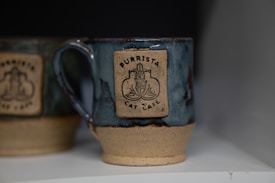 A ceramic mug with a blue glaze and a beige bottom adorned with a logo for 'Purrista Cat Cafe' featuring a stylized cat design. Another mug with similar features is partially visible in the background.