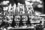 Intricate metallic components of a musical instrument, possibly a brass instrument like a trumpet or French horn, are visible. The image focuses on the decorative engravings and the complex network of valves and tubing, captured in a soft black and white color scheme with a bokeh effect in the background.