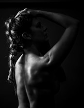 A silhouette of a person in a side profile, with one hand placed on the head, showcasing a muscular back and braided hair. The image is in black and white, accentuating shadows and highlights across the skin.