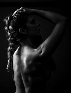 A silhouette of a person in a side profile, with one hand placed on the head, showcasing a muscular back and braided hair. The image is in black and white, accentuating shadows and highlights across the skin.