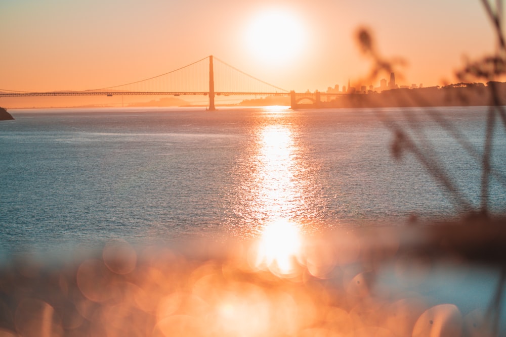 the sun is setting over the water with a bridge in the background