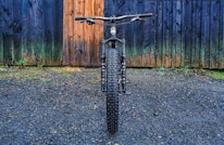 A mountain bike with thick tires is positioned on a gravel surface, directly facing the viewer. The backdrop features a rustic, multi-colored wooden wall with hues of blue and brown, adding texture to the scene.