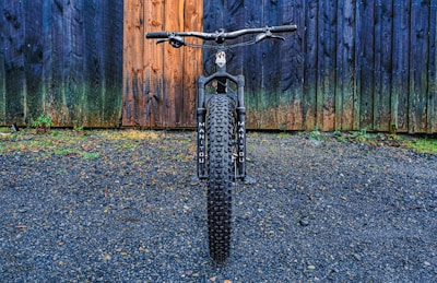 A mountain bike with thick tires is positioned on a gravel surface, directly facing the viewer. The backdrop features a rustic, multi-colored wooden wall with hues of blue and brown, adding texture to the scene.