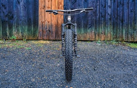 A mountain bike with thick tires is positioned on a gravel surface, directly facing the viewer. The backdrop features a rustic, multi-colored wooden wall with hues of blue and brown, adding texture to the scene.