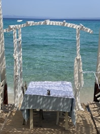 A serene seaside setting featuring a table with a white tablecloth near the water. The area is shaded by a wooden pergola casting patterned shadows on the table. The calm, clear blue sea is visible in the background, with a small white boat floating in the distance.