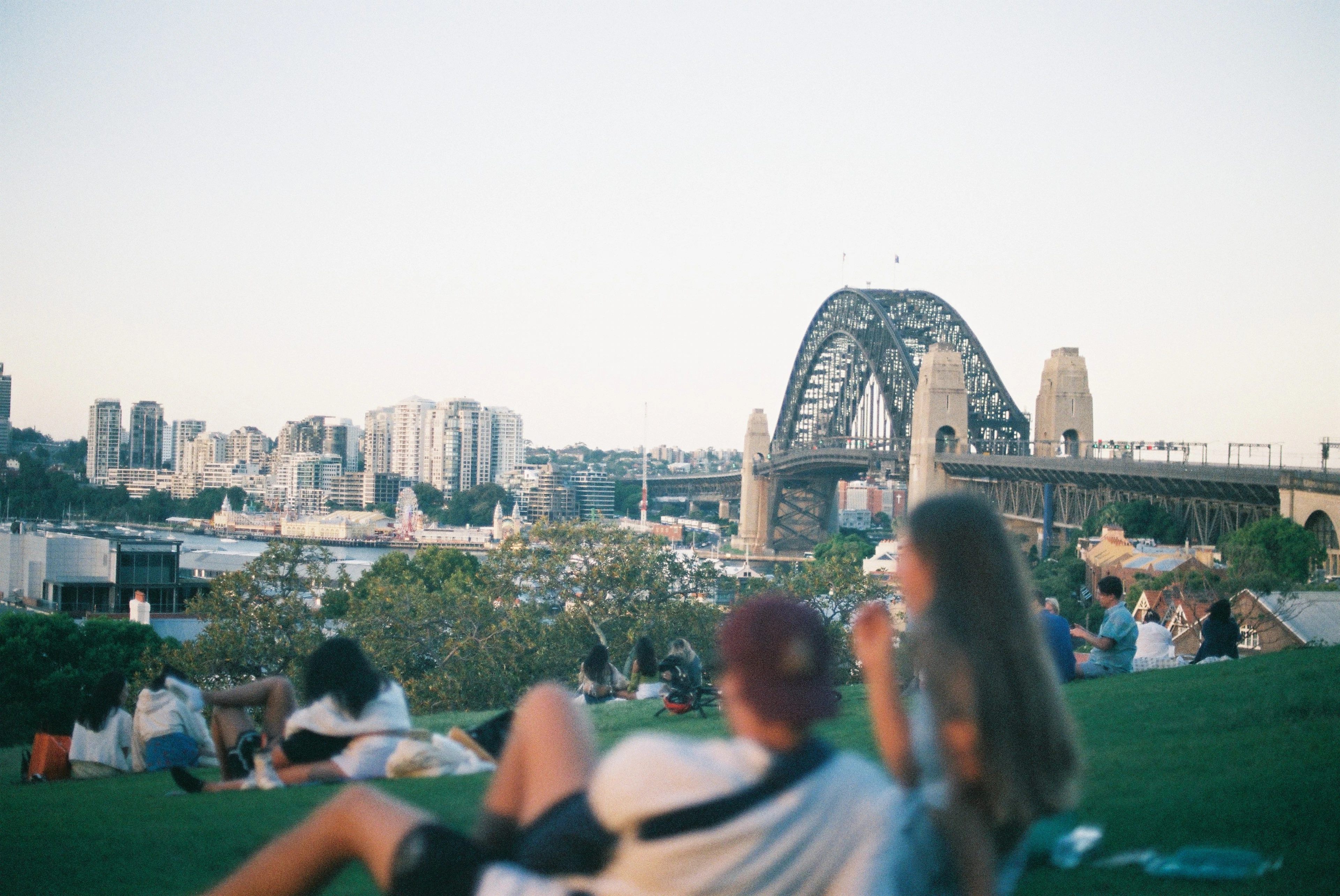 People sit on a grassy lawn watching the Sydney Harbour Bridge glow at sunset