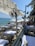 A beachfront restaurant with tables covered in white tablecloths and surrounded by wooden chairs. There is a view of the sea with people swimming and sunbathing on a sunny day. The area is shaded by a white fabric canopy, with fans mounted on poles. Palm trees line the right side.