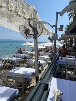 A beachfront restaurant with tables covered in white tablecloths and surrounded by wooden chairs. There is a view of the sea with people swimming and sunbathing on a sunny day. The area is shaded by a white fabric canopy, with fans mounted on poles. Palm trees line the right side.