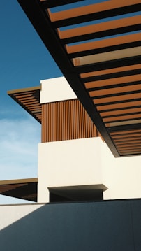 A modern architectural structure featuring clean lines and geometric shapes. The design incorporates horizontal wooden slats and a prominent white facade set against a clear blue sky, with contrasting shadows creating a sense of depth.