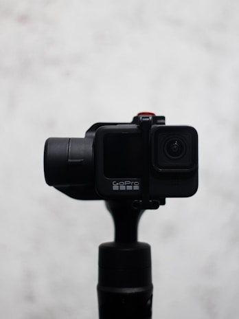 A sleek, black camera mounted on a gimbal with a minimalist design. The camera features a small lens on the front and a noticeable brand logo. The background is a soft, blurred white which contrasts with the camera, making it the focal point.
