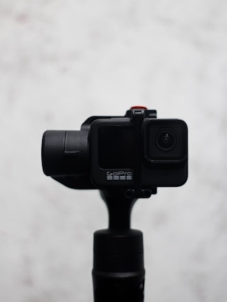A sleek, black camera mounted on a gimbal with a minimalist design. The camera features a small lens on the front and a noticeable brand logo. The background is a soft, blurred white which contrasts with the camera, making it the focal point.