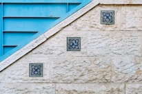 A textured stone wall features three decorative blue tiles arranged diagonally along its surface. Adjacent to the wall is a blue metallic structure with horizontal slats, creating a geometric contrast with the stone. The tiles display intricate floral patterns in shades of blue and green.