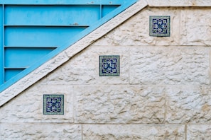 A textured stone wall features three decorative blue tiles arranged diagonally along its surface. Adjacent to the wall is a blue metallic structure with horizontal slats, creating a geometric contrast with the stone. The tiles display intricate floral patterns in shades of blue and green.