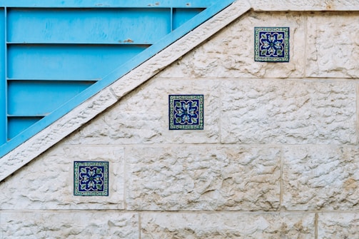 A textured stone wall features three decorative blue tiles arranged diagonally along its surface. Adjacent to the wall is a blue metallic structure with horizontal slats, creating a geometric contrast with the stone. The tiles display intricate floral patterns in shades of blue and green.