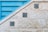 A textured stone wall features three decorative blue tiles arranged diagonally along its surface. Adjacent to the wall is a blue metallic structure with horizontal slats, creating a geometric contrast with the stone. The tiles display intricate floral patterns in shades of blue and green.