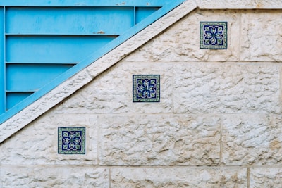 A textured stone wall features three decorative blue tiles arranged diagonally along its surface. Adjacent to the wall is a blue metallic structure with horizontal slats, creating a geometric contrast with the stone. The tiles display intricate floral patterns in shades of blue and green.