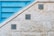 A textured stone wall features three decorative blue tiles arranged diagonally along its surface. Adjacent to the wall is a blue metallic structure with horizontal slats, creating a geometric contrast with the stone. The tiles display intricate floral patterns in shades of blue and green.