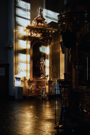 A dimly lit interior with intricately designed gilded furniture and decor, accentuated by the warm glow of sunlight streaming in, casting shadows across the polished floor. The ornate elements include candles and elaborate carvings, creating a sense of historical grandeur.