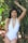 A woman stands confidently amidst a backdrop of lush greenery, wearing a white swimsuit with lace-up sides. Her long dark hair cascades down, and she accessorizes with long earrings, a bracelet, and anklets. The setting appears to be a garden or a forested area with bamboo leaves surrounding her.