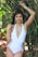 A woman stands confidently amidst a backdrop of lush greenery, wearing a white swimsuit with lace-up sides. Her long dark hair cascades down, and she accessorizes with long earrings, a bracelet, and anklets. The setting appears to be a garden or a forested area with bamboo leaves surrounding her.