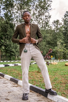 A young man stands confidently on a paved pathway, surrounded by greenery. He is wearing an open brown blazer, revealing a cross necklace and bare chest, along with light-colored trousers and black shoes.
