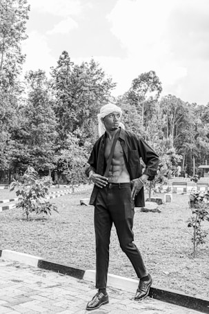 A person stands confidently in an outdoor setting, dressed in a fashionable black outfit with an open shirt revealing a muscular torso. The background features a garden with trees and shrubs, lined with small paths.