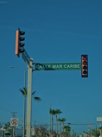 A street intersection with a traffic light displaying a red signal. There is a green street sign reading 'CALLE MAR CARIBE' mounted on the same pole as the traffic light. In the background, there are palm trees and power lines, with clear blue skies above.