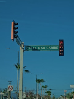 A street intersection with a traffic light displaying a red signal. There is a green street sign reading 'CALLE MAR CARIBE' mounted on the same pole as the traffic light. In the background, there are palm trees and power lines, with clear blue skies above.