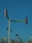A street intersection with a traffic light displaying a red signal. There is a green street sign reading 'CALLE MAR CARIBE' mounted on the same pole as the traffic light. In the background, there are palm trees and power lines, with clear blue skies above.