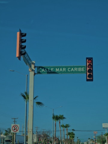 A street intersection with a traffic light displaying a red signal. There is a green street sign reading 'CALLE MAR CARIBE' mounted on the same pole as the traffic light. In the background, there are palm trees and power lines, with clear blue skies above.