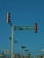 A street intersection with a traffic light displaying a red signal. There is a green street sign reading 'CALLE MAR CARIBE' mounted on the same pole as the traffic light. In the background, there are palm trees and power lines, with clear blue skies above.