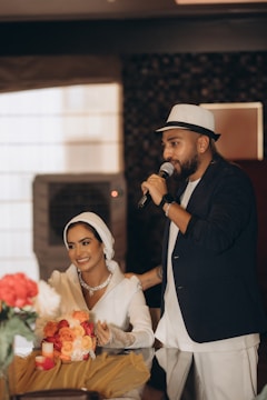 A man wearing a hat and holding a microphone stands beside a smiling woman dressed in white. The woman is seated at a table with a bouquet of red and peach roses, along with candles.