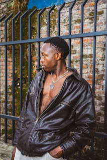 A person wearing a dark leather jacket stands in front of an iron fence with a brick wall background, partially covered in some greenery. The individual is looking to the side, wearing a necklace with a noticeable pendant, and the jacket is slightly open, revealing a part of their chest.