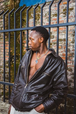 A person wearing a dark leather jacket stands in front of an iron fence with a brick wall background, partially covered in some greenery. The individual is looking to the side, wearing a necklace with a noticeable pendant, and the jacket is slightly open, revealing a part of their chest.