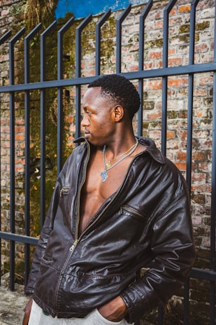 A person wearing a dark leather jacket stands in front of an iron fence with a brick wall background, partially covered in some greenery. The individual is looking to the side, wearing a necklace with a noticeable pendant, and the jacket is slightly open, revealing a part of their chest.