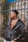 A person wearing a dark leather jacket stands in front of an iron fence with a brick wall background, partially covered in some greenery. The individual is looking to the side, wearing a necklace with a noticeable pendant, and the jacket is slightly open, revealing a part of their chest.