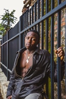 A person in a black leather jacket leans against a metal fence. They wear a necklace with a pendant shaped like the African continent. The background includes an old brick wall partly covered in moss and some greenery.