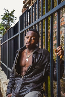 A person in a black leather jacket leans against a metal fence. They wear a necklace with a pendant shaped like the African continent. The background includes an old brick wall partly covered in moss and some greenery.
