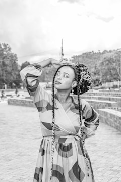 A person with long braided hair is standing outdoors with one hand resting on their head and the other on their hip. They are wearing a patterned dress, and the background features steps made of bricks and trees. The image is in black and white, giving it a classic look.