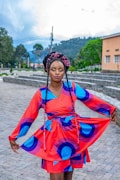 A person stands outdoors wearing a vibrant, colorful dress with a geometric pattern featuring blue and purple arcs on a red background. She is holding the sides of the dress out slightly while looking directly at the camera. Her hair is styled in braids with pink highlights. The background includes a paved area, some greenery, and buildings with a cloudy sky above.