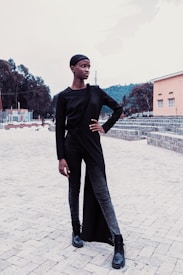 A person in stylish black attire poses confidently on a paved area. The outfit features a long black overcoat with a unique asymmetrical design, paired with black jeans and boots. The background includes trees and a pastel-colored building.