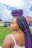 A woman with large, braided hair featuring black and purple twists stands outdoors. She is wearing a white top and gold hoop earrings. The background includes a grassy area and buildings under a partly cloudy sky.