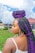 A woman with large, braided hair featuring black and purple twists stands outdoors. She is wearing a white top and gold hoop earrings. The background includes a grassy area and buildings under a partly cloudy sky.