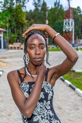 A person with braided hair poses outdoors, hand resting on their head, wearing a black floral dress and pearl necklace, with a scenic background featuring trees and a metal tower.